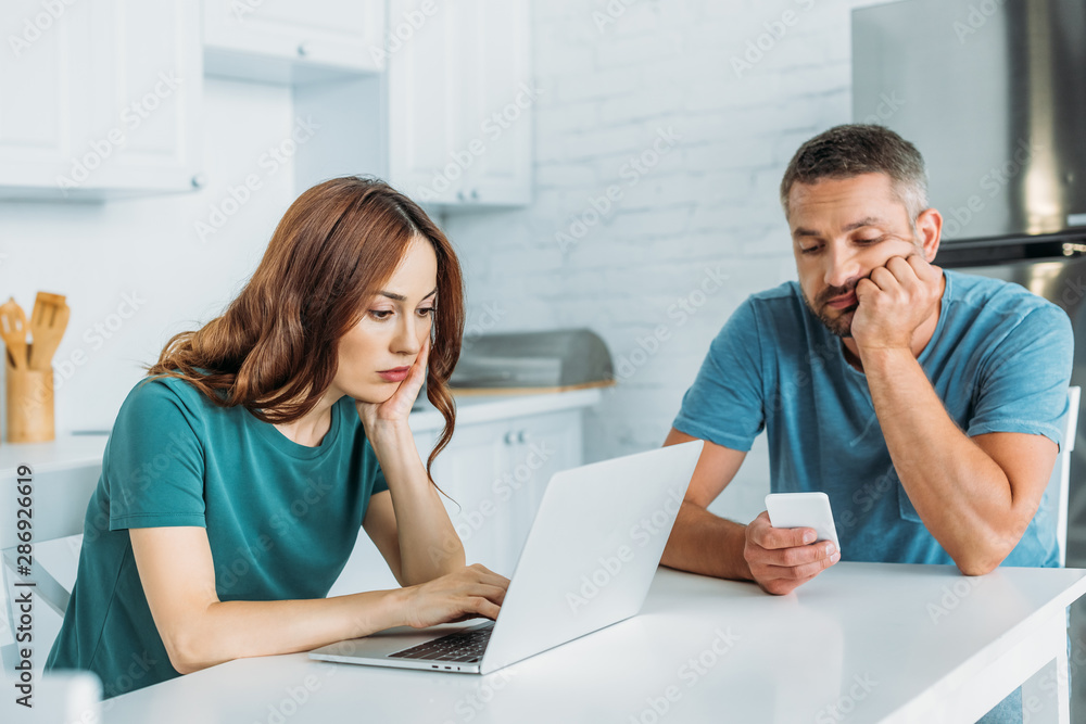 thoughtful man using smartphone while sitting at kitchen table near wife using laptop thoughtful man using smartphone while sitting at kitchen table near wife using laptop