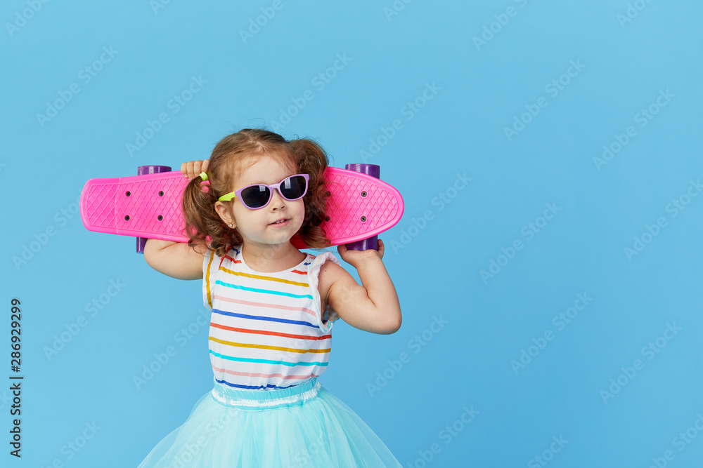 Cool little toddler girl in bright clothes and sunglasses carrying a skateboard over his shoulder and looking at the camera isolated on blue background