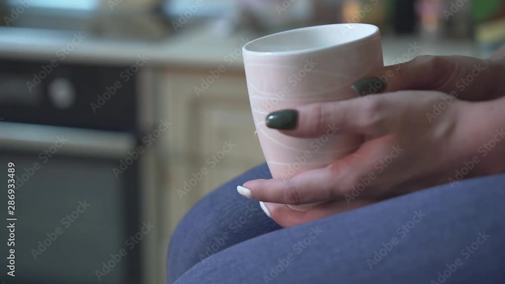 Close-up of girl's hands in the kitchen. The girl holds a white mug in her hands in the kitchen against the background of the kitchen setting. Close-up of girl's hands in the kitchen. The girl holds a white mug in her hands in the kitchen against the background of the kitchen setting.