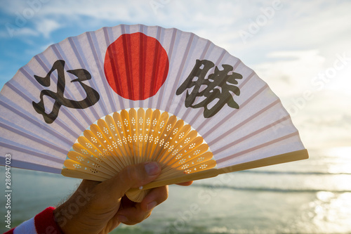 Hand of Japanese sports supporter holding a fan decorated with kanji characters spelling out hissho (English translation: certain victory) over a sunrise sea horizon photo