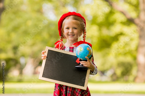 little blonde first grader girl in red dress and beret holding an empty drawing Board and globe © КРИСТИНА Игумнова