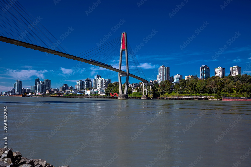 Fototapeta premium Sky-train bridge linking Surrey and New Westminster over the Fraser River, city on the opposite bank, blue sky with white clouds on a background 