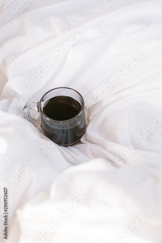 Cup of black coffee in clear glass mug on white comforter in bed, coffee in bed, copy space, cozy mornings