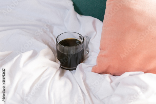 Cup of black coffee in clear glass mug on white comforter next to pink and teal pillows in bed, room service, copy space, cozy mornings