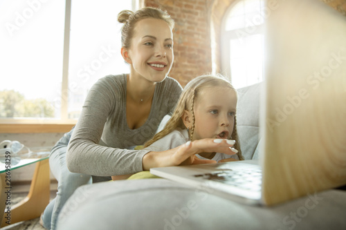 Female caucasian teacher and little girl, or mom and daughter. Homeschooling. Sitting on the sofa and using laptop for knowledges getting while lesson is. Education, school, studying concept.