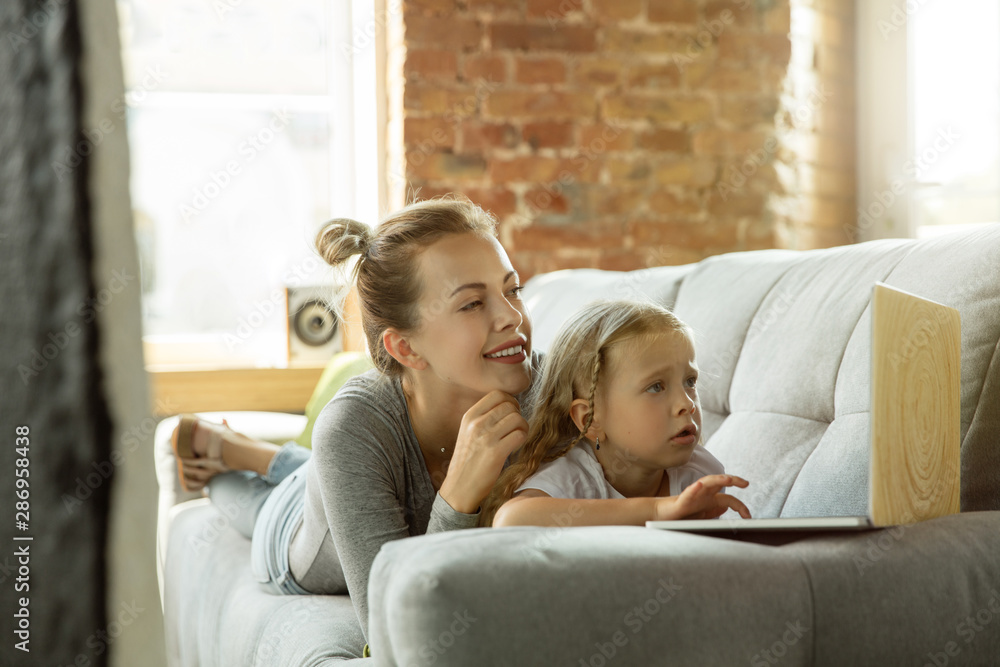 Female caucasian teacher and little girl, or mom and daughter. Homeschooling. Lying on the sofa and using laptop for knowledges getting while lesson is. Education, school, studying concept. Female caucasian teacher and little girl, or mom and daughter. Homeschooling. Lying on the sofa and using laptop for knowledges getting while lesson is. Education, school, studying concept.