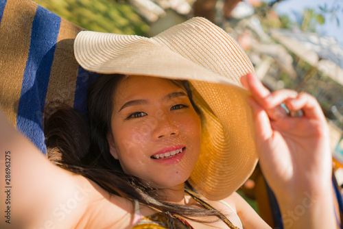 young beautiful and happy tourist Asian Korean woman in Summer hat taking selfie at tropical resort pool hammock smiling carefree and natural in holidays getaway photo