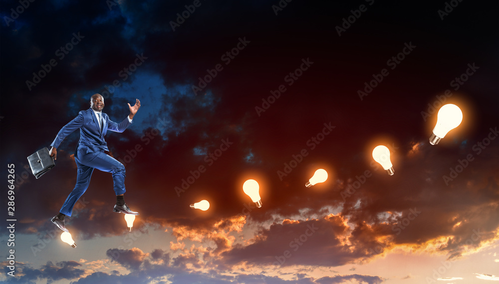 Black businessman running on hypothetical stairs made of burning lamps on a dark cloudy evening sky background