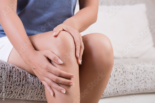 Closeup young woman sitting on sofa and feeling knee pain and she massage her knee at home. Healthcare and medical concept.