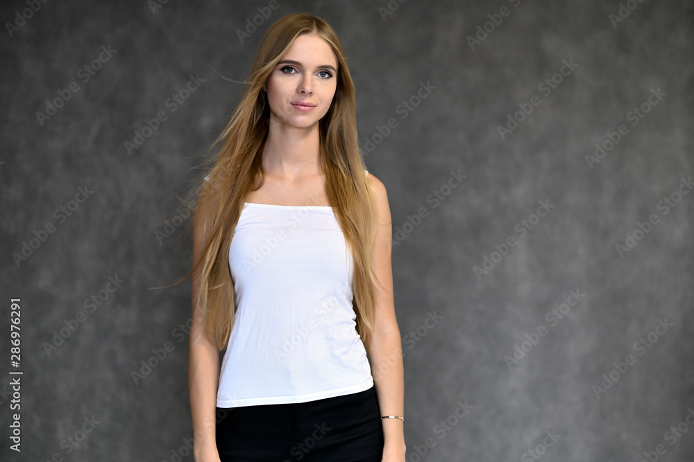 Portrait of a beautiful pretty girl student in a white T-shirt with long curly hair on a gray background. Beauty and brightness. Shows hands to the side, smiles with different emotions. Portrait of a beautiful pretty girl student in a white T-shirt with long curly hair on a gray background. Beauty and brightness. Shows hands to the side, smiles with different emotions.