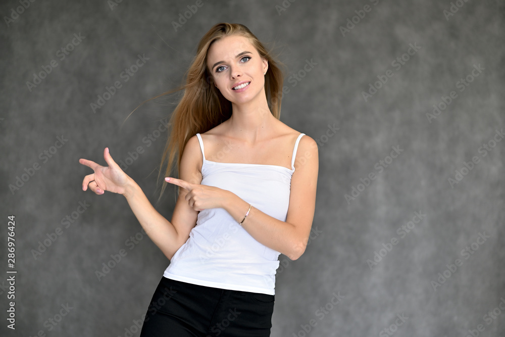 Portrait of a beautiful pretty girl student in a white T-shirt with long curly hair on a gray background. Beauty and brightness. Shows hands to the side, smiles with different emotions. Portrait of a beautiful pretty girl student in a white T-shirt with long curly hair on a gray background. Beauty and brightness. Shows hands to the side, smiles with different emotions.