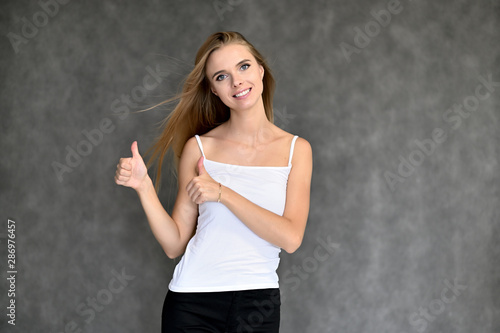 Portrait of a beautiful pretty girl student in a white T-shirt with long curly hair on a gray background. Beauty and brightness. Shows hands to the side, smiles with different emotions.