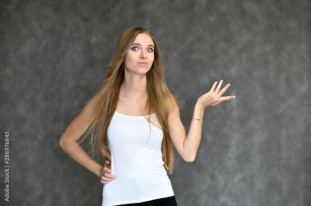 Portrait of a beautiful pretty girl student in a white T-shirt with long curly hair on a gray background. Beauty and brightness. Shows hands to the side, smiles with different emotions. Portrait of a beautiful pretty girl student in a white T-shirt with long curly hair on a gray background. Beauty and brightness. Shows hands to the side, smiles with different emotions.