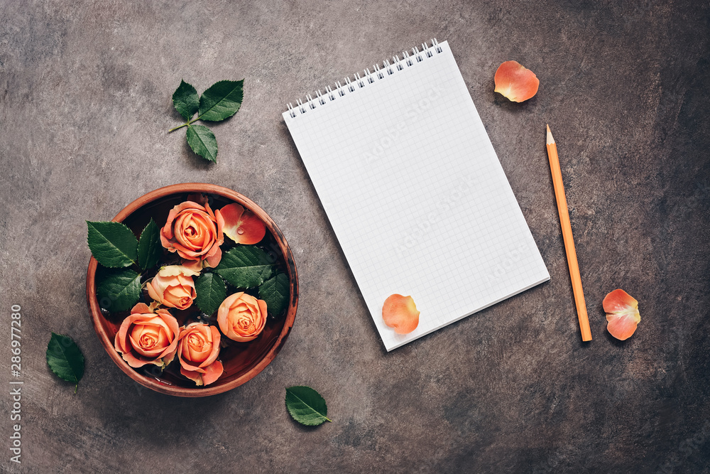 Blank notepad, ceramic bowl with flowers of coral roses and pencil on a dark rustic background.. Top view, flat lay feminine beautiful background. Blank notepad, ceramic bowl with flowers of coral roses and pencil on a dark rustic background.. Top view, flat lay feminine beautiful background.