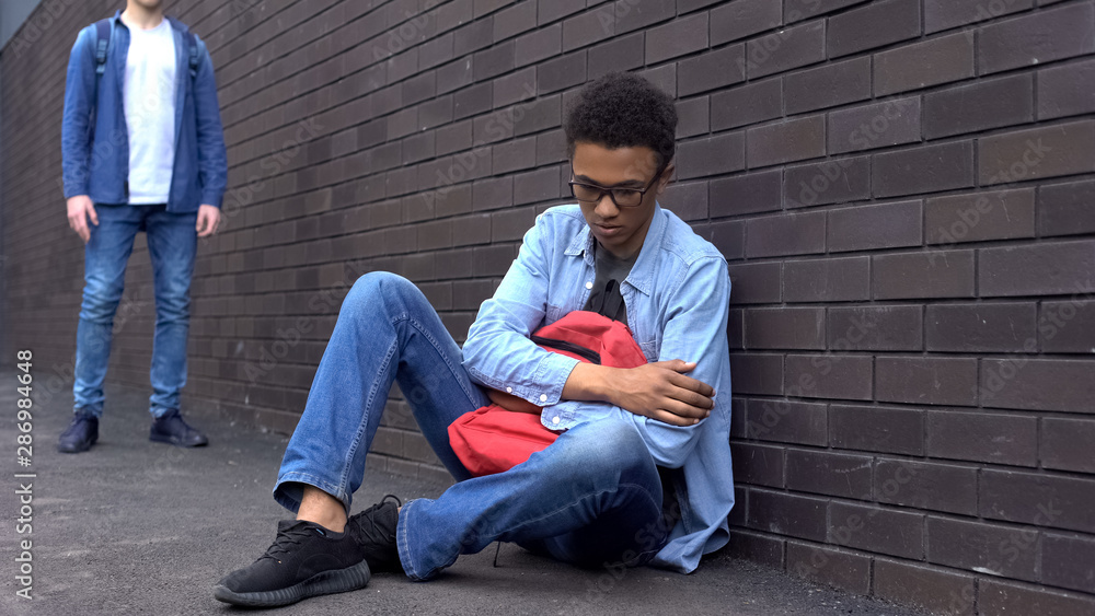 Bullied afro-american schoolboy sitting on ground, scared of offender behind