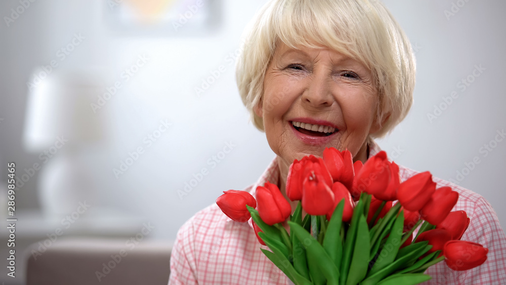 Adorable elderly female holding bunch of tulips and smiling at camera, present Adorable elderly female holding bunch of tulips and smiling at camera, present