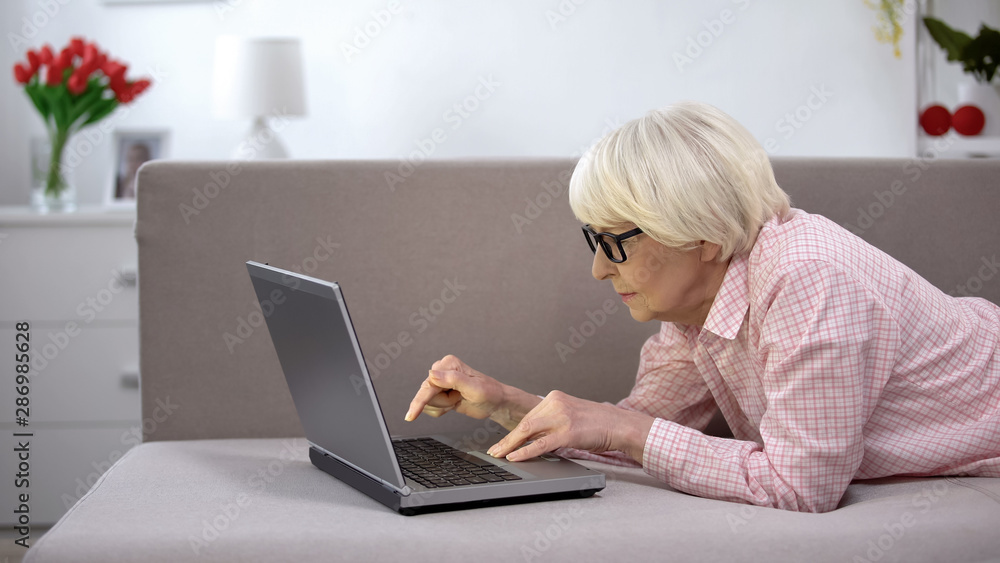 Old woman in eyeglasses attentively typing on laptop, app for elderly people Old woman in eyeglasses attentively typing on laptop, app for elderly people