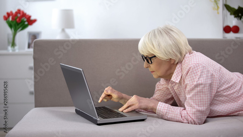 Old woman in eyeglasses attentively typing on laptop, app for elderly people