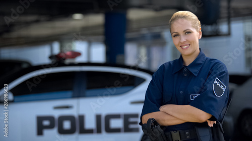 Bild auf Leinwand Woman police officer smiling, standing near patrol car with crossed hands, law