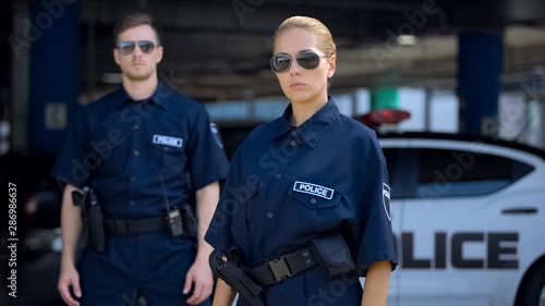 Serious woman and man police officers in sunglasses standing outdoor, safety