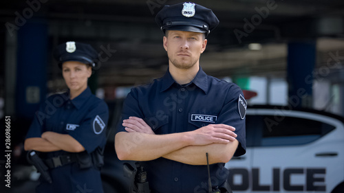 Obraz na plátně Confident male and female police officers in uniform standing near patrol car