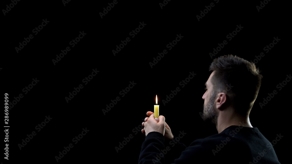 Fototapeta premium Praying man holding burning candle on dark background, spiritual ritual, faith