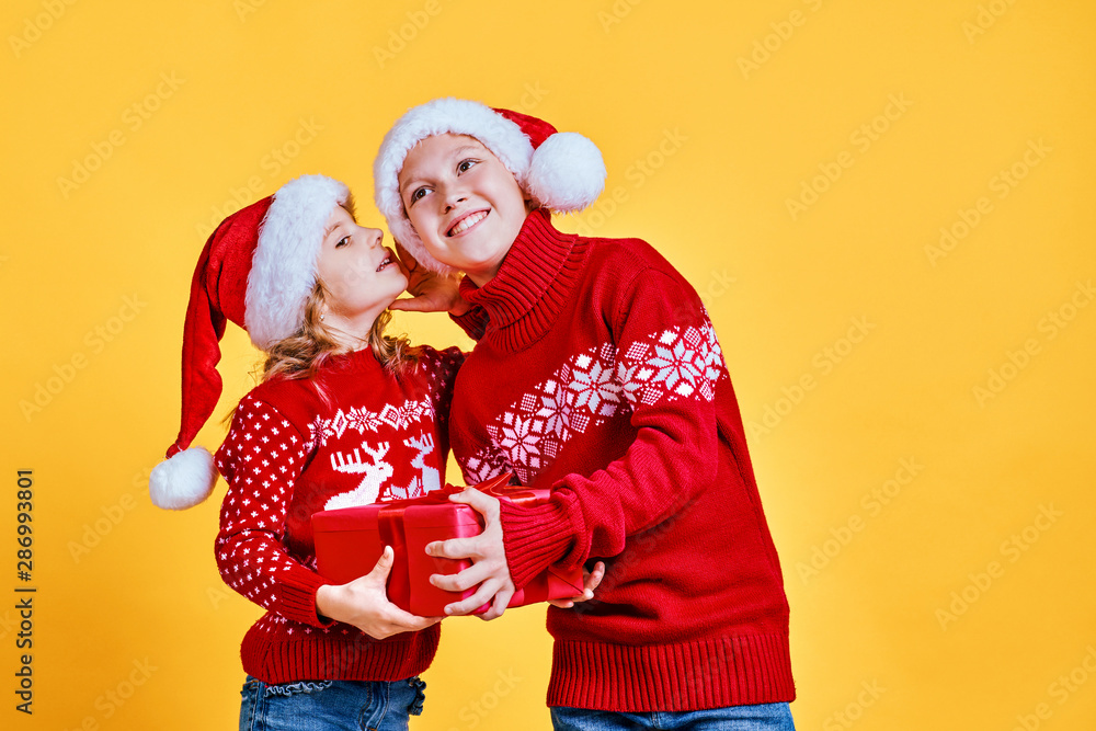 Fototapeta premium Little girl in red sweater with deer whispering secret to cheerful boy holding gift box on Christmas against yellow background