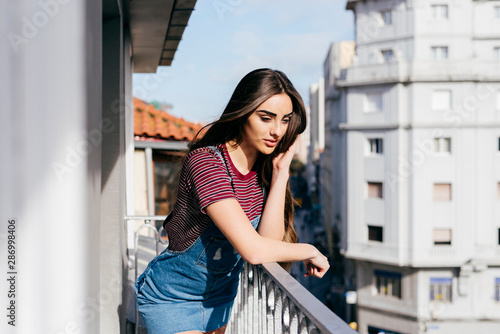 Cute brunette girl leaning on balcony and looking at the street at home on a sunny day © Manuel
