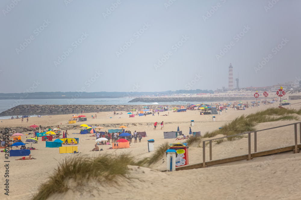View of pedestrian wooden path on beach, people walking and people taking sunbath on beach, lighthouse as background