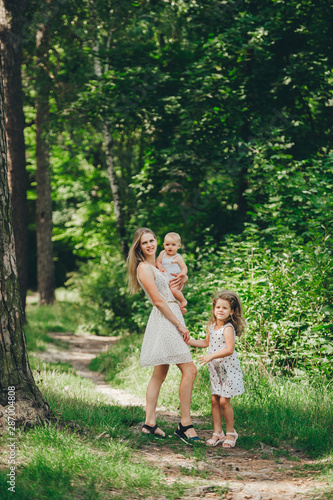 Mother and two little daughter in summer dresses go through the woods in the summer. Mother and children outdoors