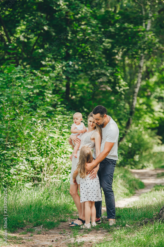Fototapeta premium Parents and two cute little daughter in holds posing on the path in a sunny day in forest