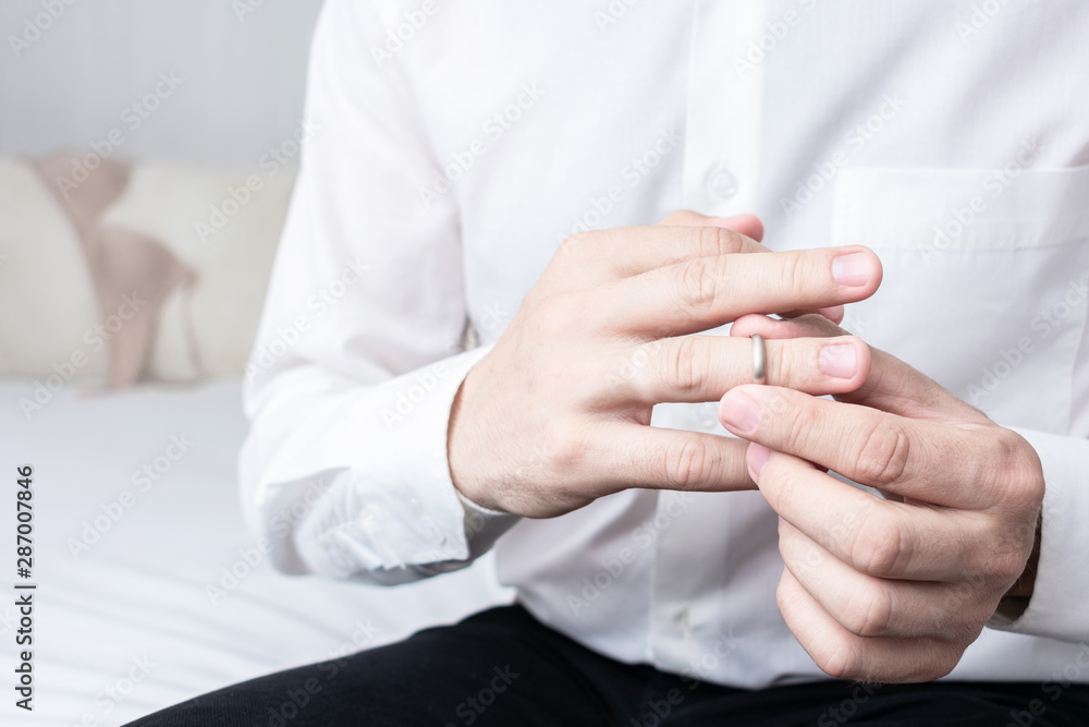 Unfaithful man taking off his wedding ring, man sitting on the bed in the hotel, his mistress's underwear in the background, closeup, cropped image, toned. The concept of infidelity in marriage