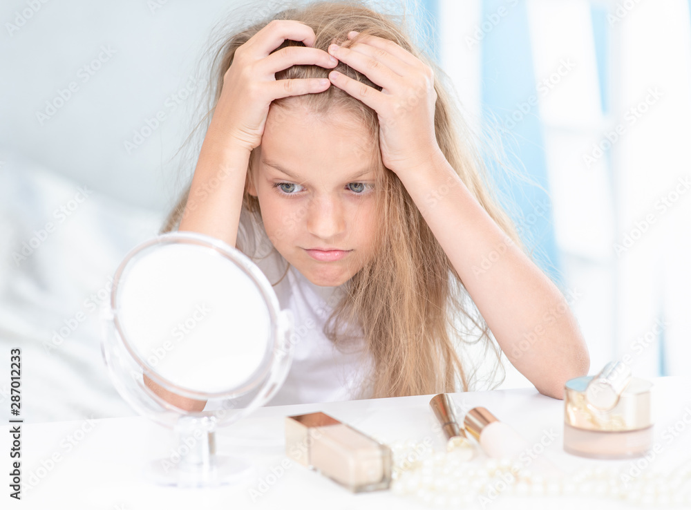 Unhappy little girl sitting and looking in the mirror at home