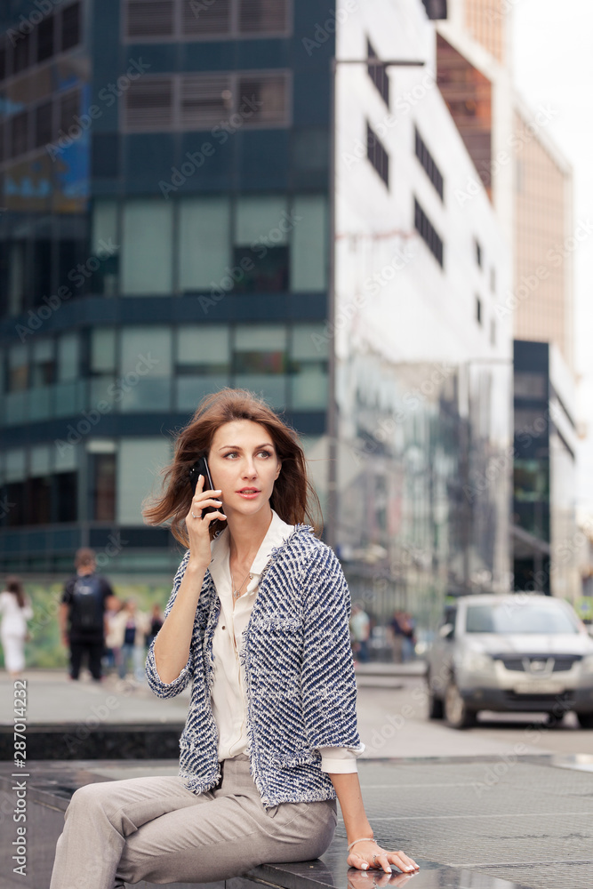 beautiful young business brunette woman dressed in elegant clothes sits on a bench on a street in the city center and talking on a cell phone. Glass building in the background