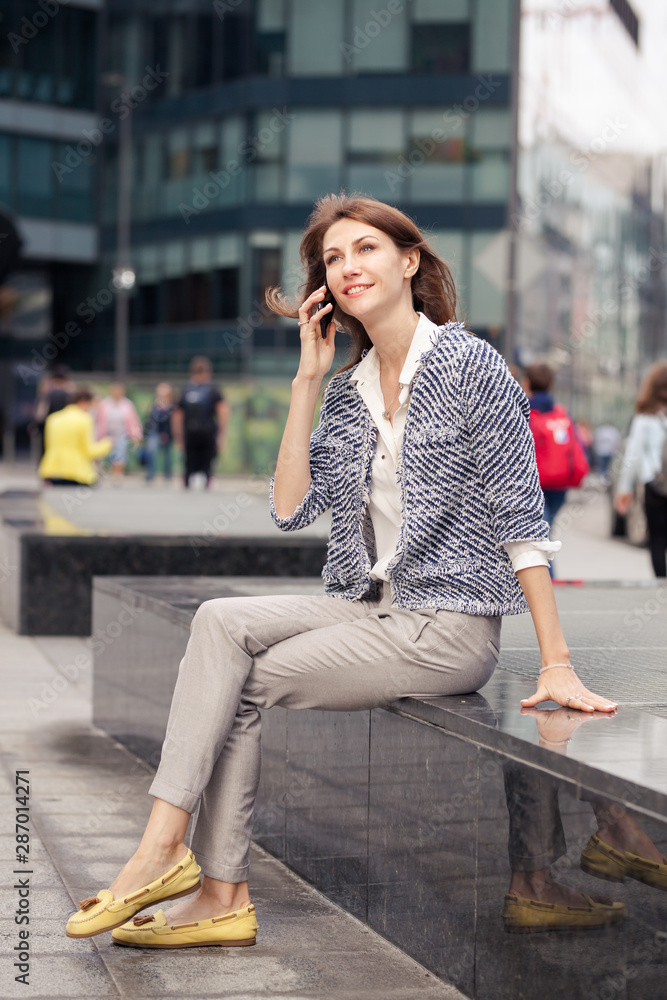 attractive smiling young business brunette girl dressed in elegant clothes sits on a bench on a street in the city center and talking on mobile phone. Glass building in the background