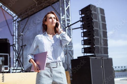 Installation of stage equipment and preparing for a live concert open air. Event manager portrait. Summer music city festival. Young serious woman stand and work with her laptop near the stage...