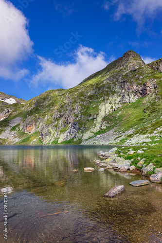 Flock of small fish, ripple in the clean, transparent water of famous Kidney lake, one of seven Rila lakes, epic, sunlit rocky summits and a blue sky with white clouds