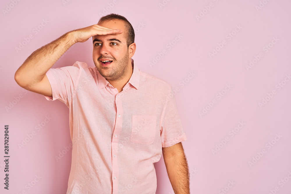 Young man wearing elegant shirt standing over isolated pink background very happy and smiling looking far away with hand over head. Searching concept. Young man wearing elegant shirt standing over isolated pink background very happy and smiling looking far away with hand over head. Searching concept.