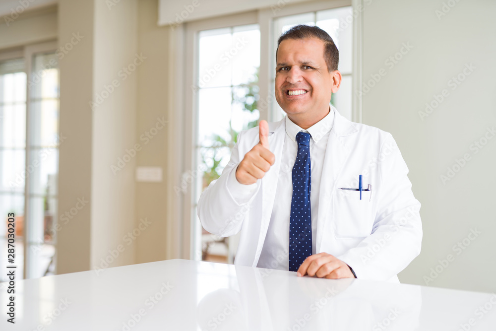 Middle age doctor man wearing medical coat at the clinic doing happy thumbs up gesture with hand. Approving expression looking at the camera showing success.
