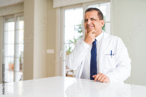 Middle age doctor man wearing medical coat at the clinic looking confident at the camera with smile with crossed arms and hand raised on chin. Thinking positive.