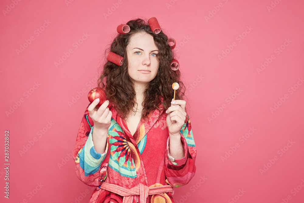 Studio shot of thoughtful pretty indecisive curly haired housewife with curlers on head feeling confused and hesitant, facing difficult choice: whether to eat healthy organic apple or unhealthy candy. Studio shot of thoughtful pretty indecisive curly haired housewife with curlers on head feeling confused and hesitant, facing difficult choice: whether to eat healthy organic apple or unhealthy candy.