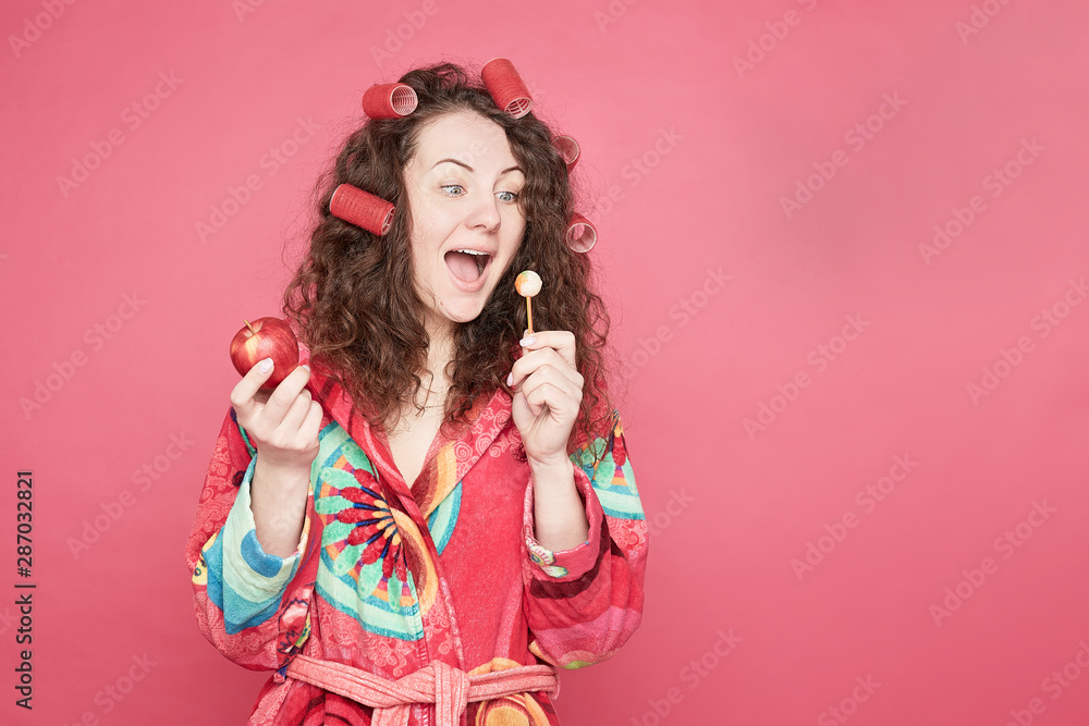Isolated on pink studio wall portrait of Caucasian blue eyed young woman in with curlers on head, feeling hungry, going to eat healthy organic apple instead of unhealthy candy. Willpower concept. Isolated on pink studio wall portrait of Caucasian blue eyed young woman in with curlers on head, feeling hungry, going to eat healthy organic apple instead of unhealthy candy. Willpower concept.