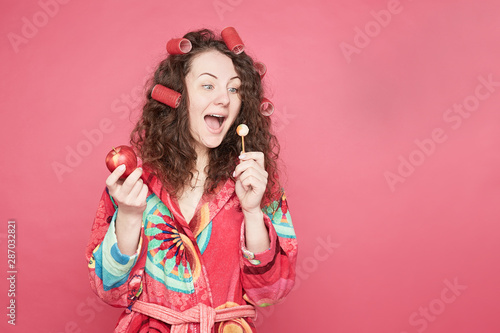 Isolated on pink studio wall portrait of Caucasian blue eyed young woman in with curlers on head, feeling hungry, going to eat healthy organic apple instead of unhealthy candy. Willpower concept.