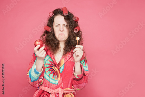 .Close up shot of grimacing housewife in colorful bathrobe feeling disgust to eat healthy organic apple or unhealthy candy, dislike to keep diet, isolated on pink studio wall with copy space.