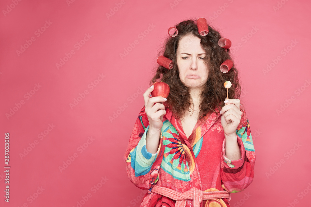 .Studio shot of pretty housewife in colorful bathrobe feeling confused and hesitant, facing difficult choice: whether to eat healthy organic apple or unhealthy candy, isolated on pink with copy space. .Studio shot of pretty housewife in colorful bathrobe feeling confused and hesitant, facing difficult choice: whether to eat healthy organic apple or unhealthy candy, isolated on pink with copy space.