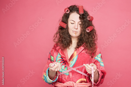 Studio close up shot of housewife wondering if she had plump out, wants to loose weight, wears colorful casual home bathrobe, can not overcome challenge, sucks candy, taking measuring tap in hands.