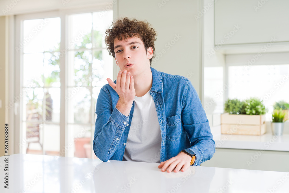 Young handsome man wearing casual denim jacket at home looking at the camera blowing a kiss with hand on air being lovely and sexy. Love expression. Young handsome man wearing casual denim jacket at home looking at the camera blowing a kiss with hand on air being lovely and sexy. Love expression.