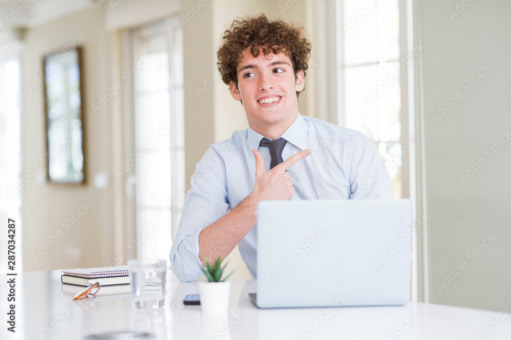 Young business man working with computer laptop at the office cheerful with a smile of face pointing with hand and finger up to the side with happy and natural expression on face