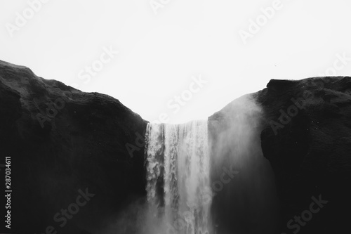 Fototapeta Naklejka Na Ścianę i Meble -  Falling water of Skogarfoss waterfall between green hills in Iceland. Gray cloudy sky and white splashes. Gothic black and white tones.