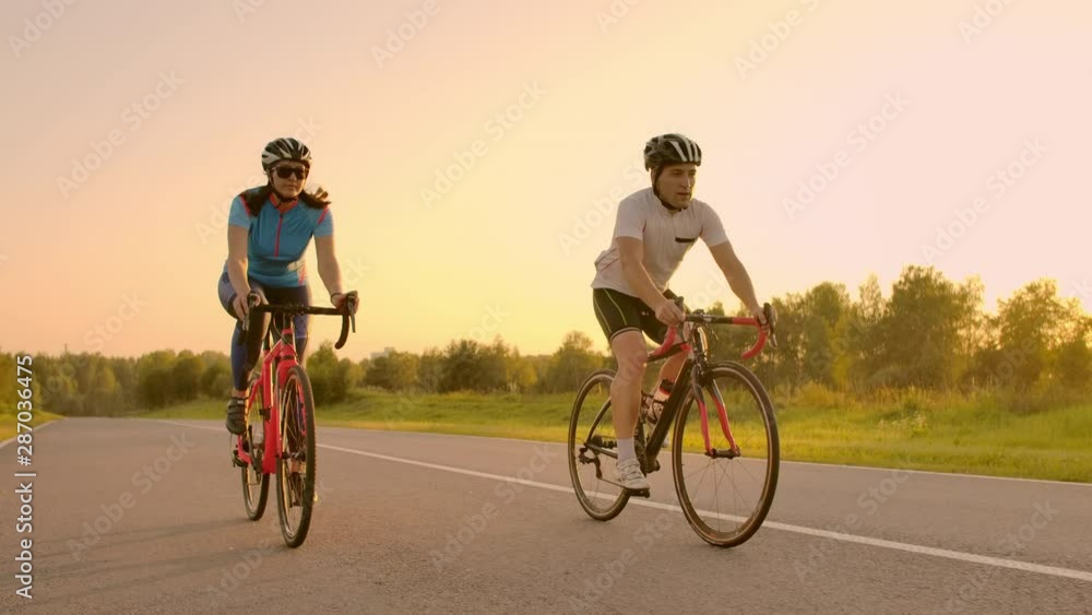 Two professional road cyclists ride their bikes on a hill. Hand held shot of two strong cyclists female and male on their training on a warm but windy summer day. Two professional road cyclists ride their bikes on a hill. Hand held shot of two strong cyclists female and male on their training on a warm but windy summer day.